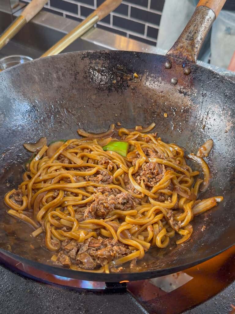 Leafy greens placed into hot wok with noodles