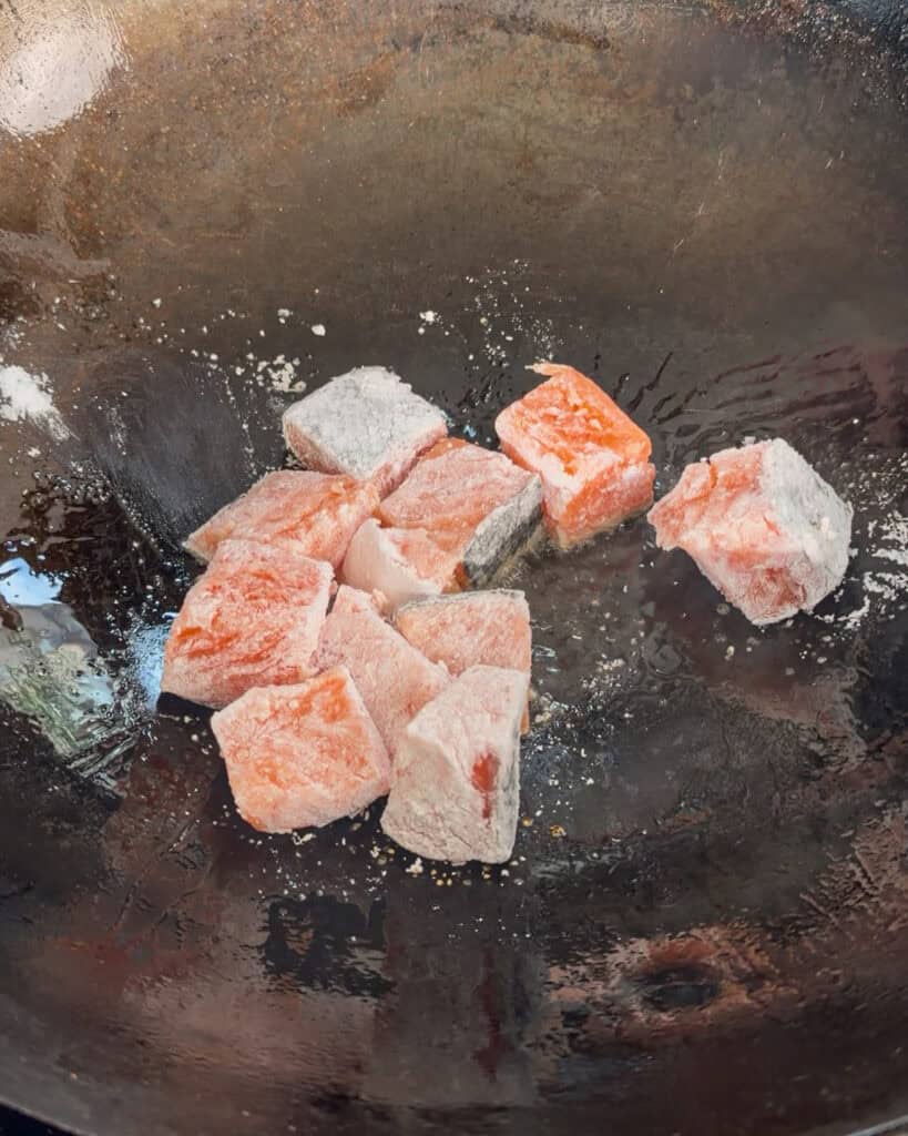 Salmon cubes are coated in white powder and placed in a wok before cooking.