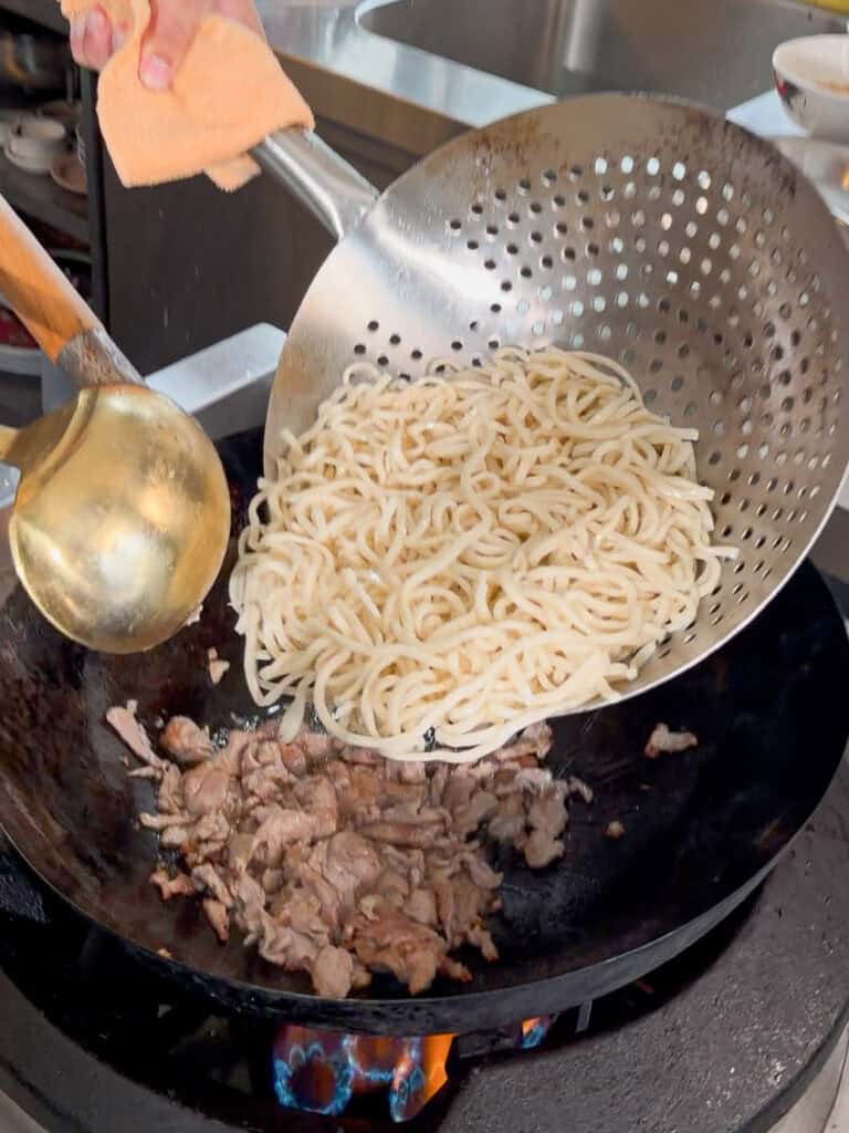 Velveted pork cooking in a hot wok as noodles are added