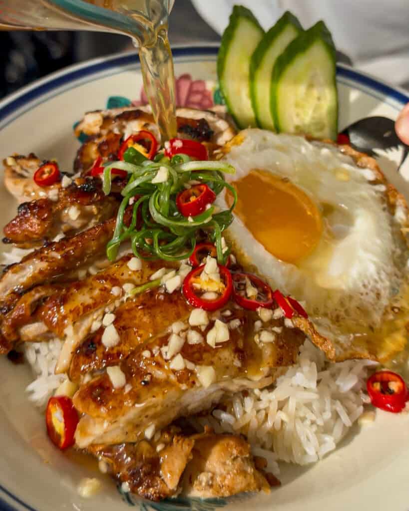 Cooked chicken served over white rice with cucumber slices and a fried egg while liquid poured