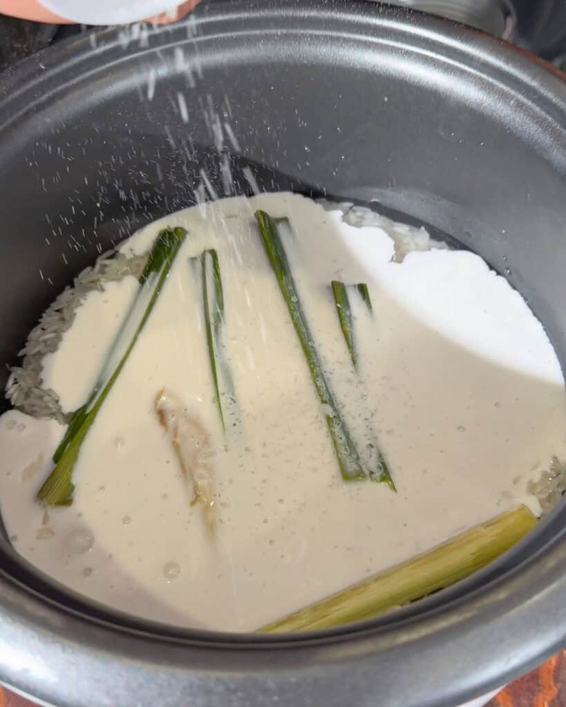 Coconut cream being poured over rice with pandan leaves in a rice cooker pot.