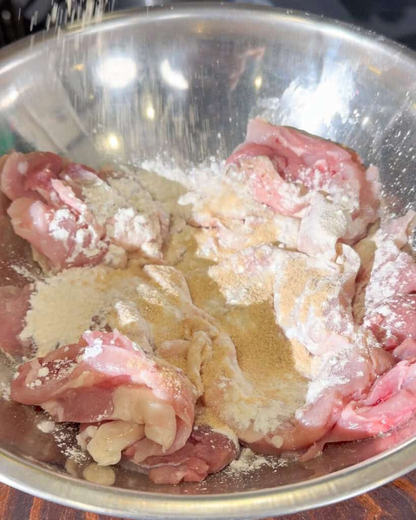 Raw chicken pieces coated with flour and seasoning powders in a metal mixing bowl.