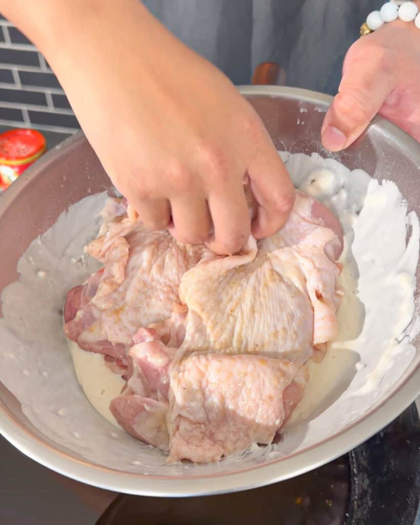Raw chicken thighs in a metal bowl with liquid seasoning being added for marinating