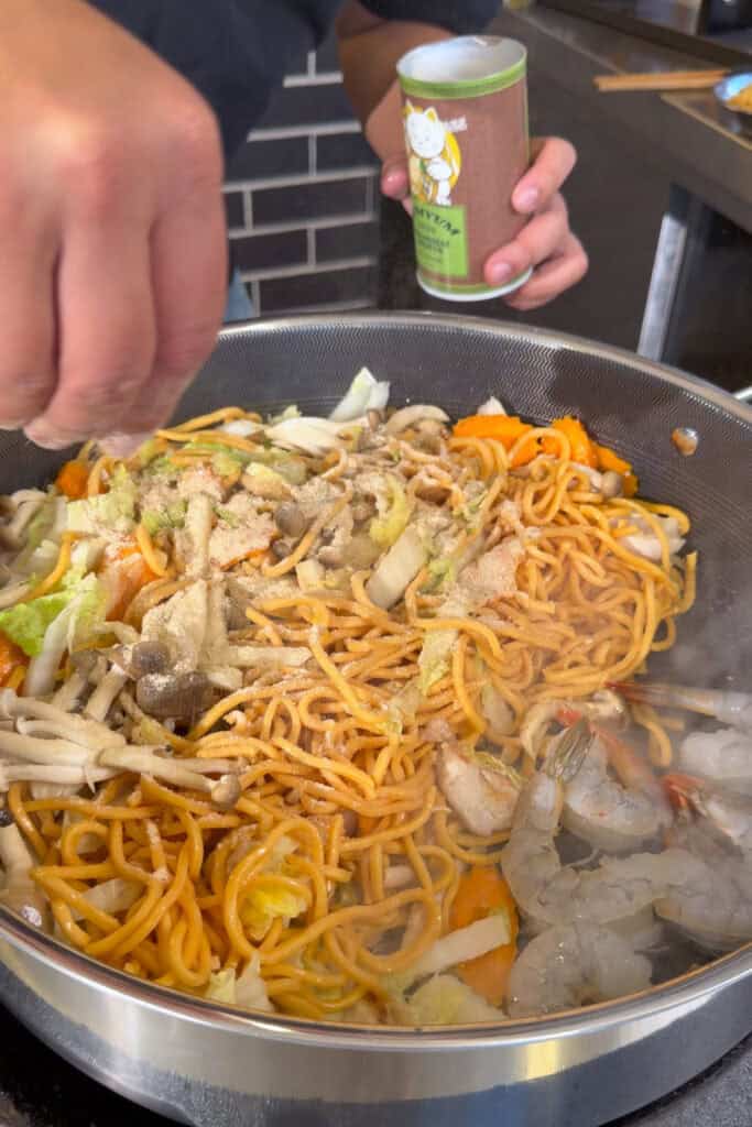 Person sprinkling seasoning over a large skillet of pasta with vegetables and mushrooms