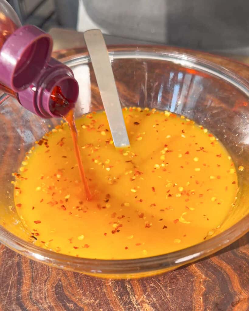 Orange spicy sauce with red pepper flakes being poured from a purple bottle into a glass bowl, with a metal spatula resting in the mix