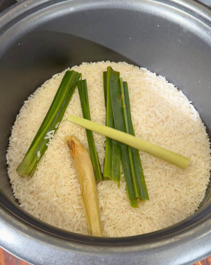 Uncooked coconut rice ingredients with pandan leaves and lemongrass in a rice cooker pot.