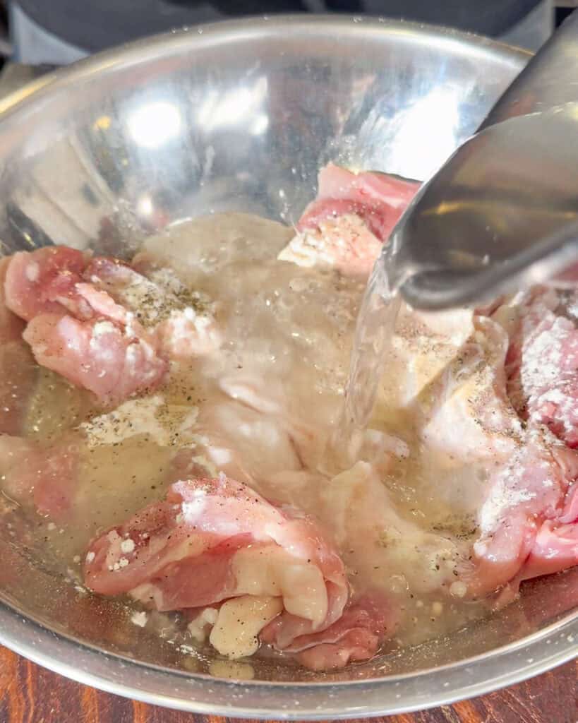 Water being poured into raw chicken with flour and black pepper in a metal bowl.