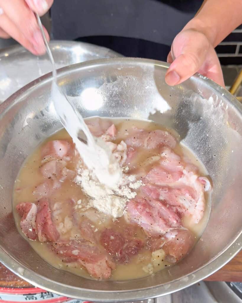 White flour mixture being added to marinated chicken pieces in a metal bowl.