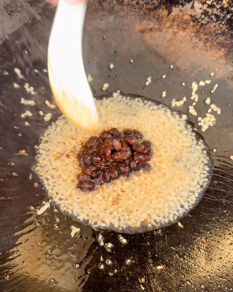 Black bean sauce added to bubbling oil and small garlic-like bits in a wok