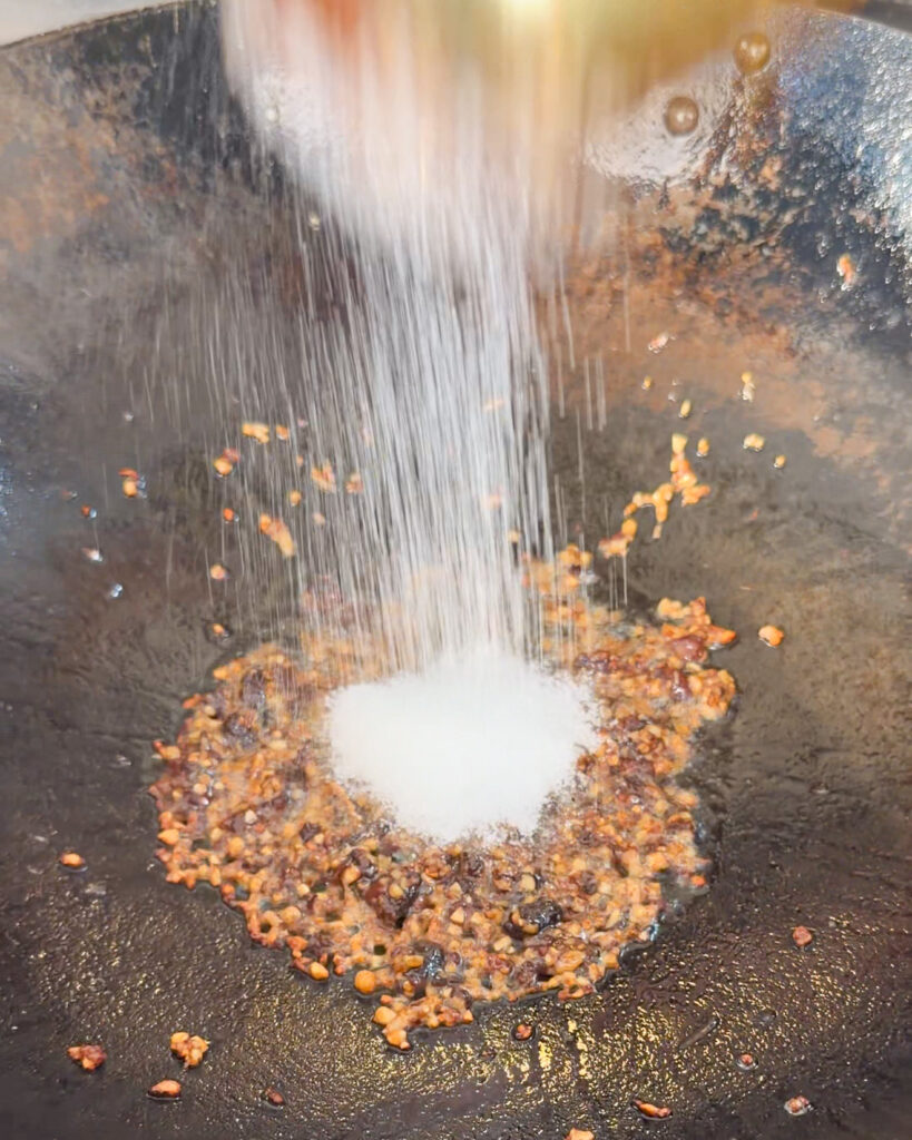 White seasoning granules added to sauce ingredients in a wok